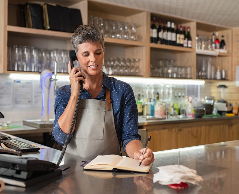 Barista woman taking orders over the phone at counter. Mature woman in grey apron writing on customer book while talking over phone in cafeteria. Waitress writing takeaway lunch while using telephone.