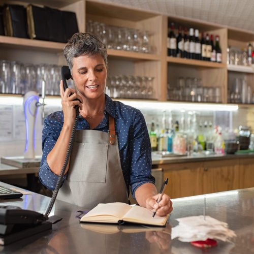 Barista woman taking orders over the phone at counter. Mature woman in grey apron writing on customer book while talking over phone in cafeteria. Waitress writing takeaway lunch while using telephone.